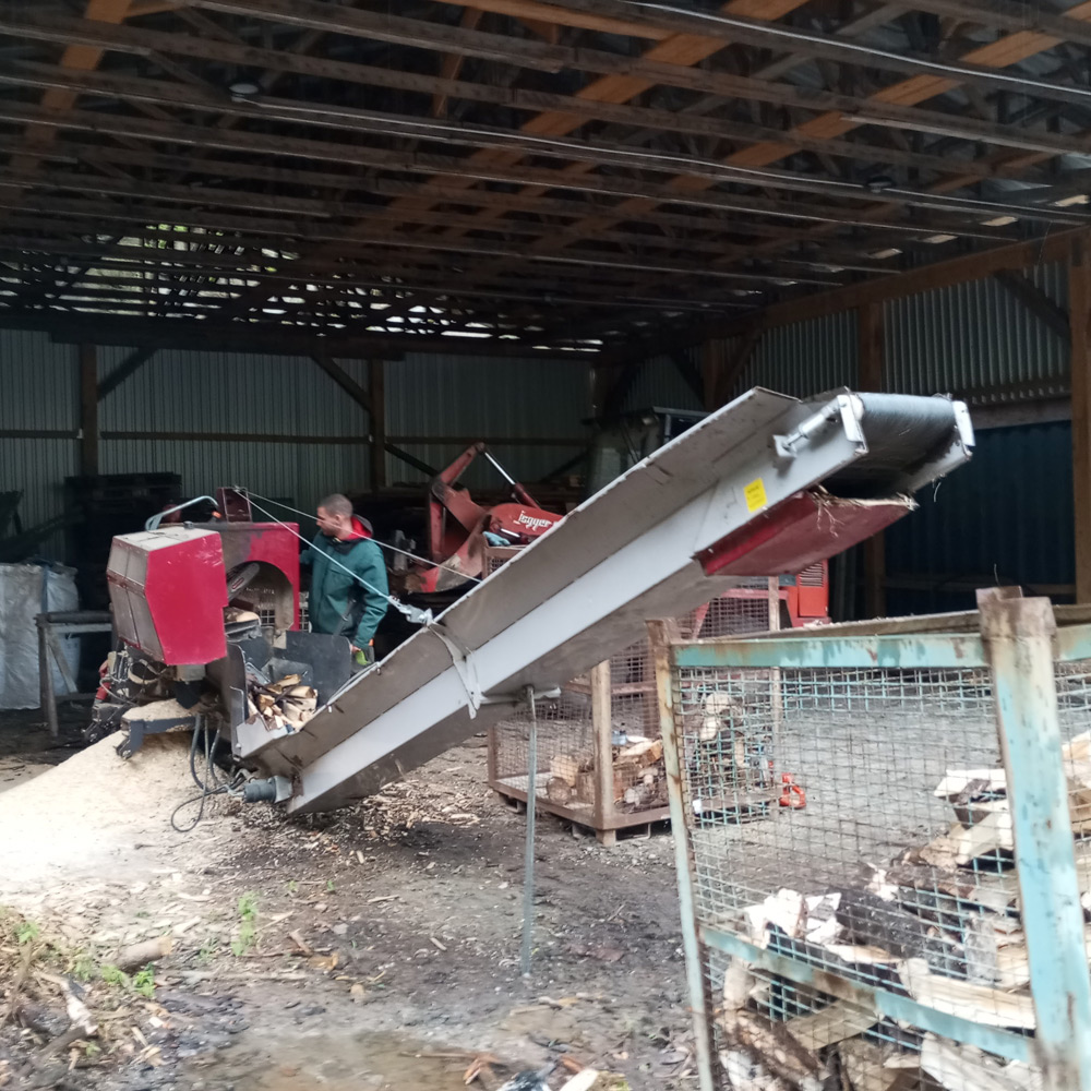 Auf dem Foto sieht man den Teilnehmer Max an einer Maschine, die das Holz spaltet. Im Vordergrund ist ein Förderband, das die Holzscheite in eine großen Drahtkorb befördert.