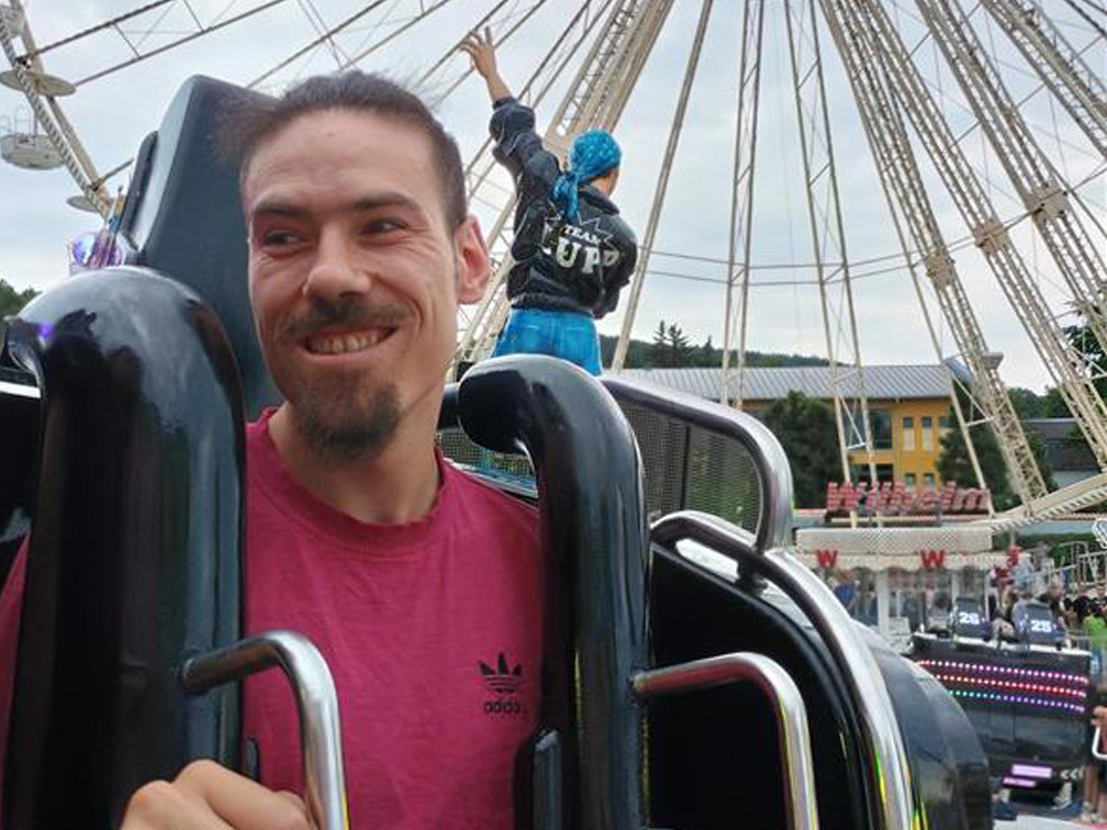 Auf dem Foto sitzt ein junger Mann in einem Fahrgeschäft. Im Hintergrund ist ein Riesenrad.