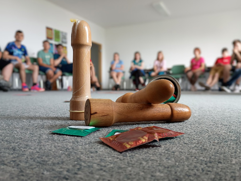 Im Vordergrund des Bildes sieht man Holz-Penis-Modelle und Kondome. Im Hintergrund ist verschwommen die Gruppe der Teilnehmenden zu sehen.